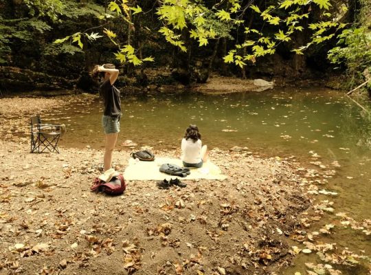 Two people relax by a quiet riverside surrounded by autumn leaves.