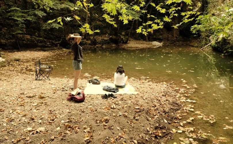 Two people relax by a quiet riverside surrounded by autumn leaves.
